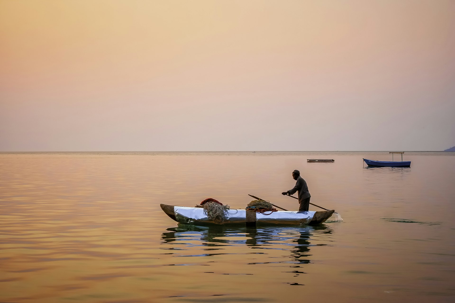 People in silhouette fishing from a canoe at sunset, with an orangey-pink sky reflected in the water. 