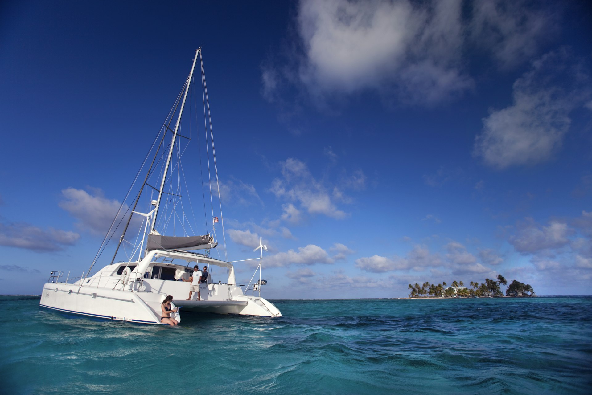 A catamaran is moored in the blue waters of Belize. There is a woman sitting on the edge of the boat. 