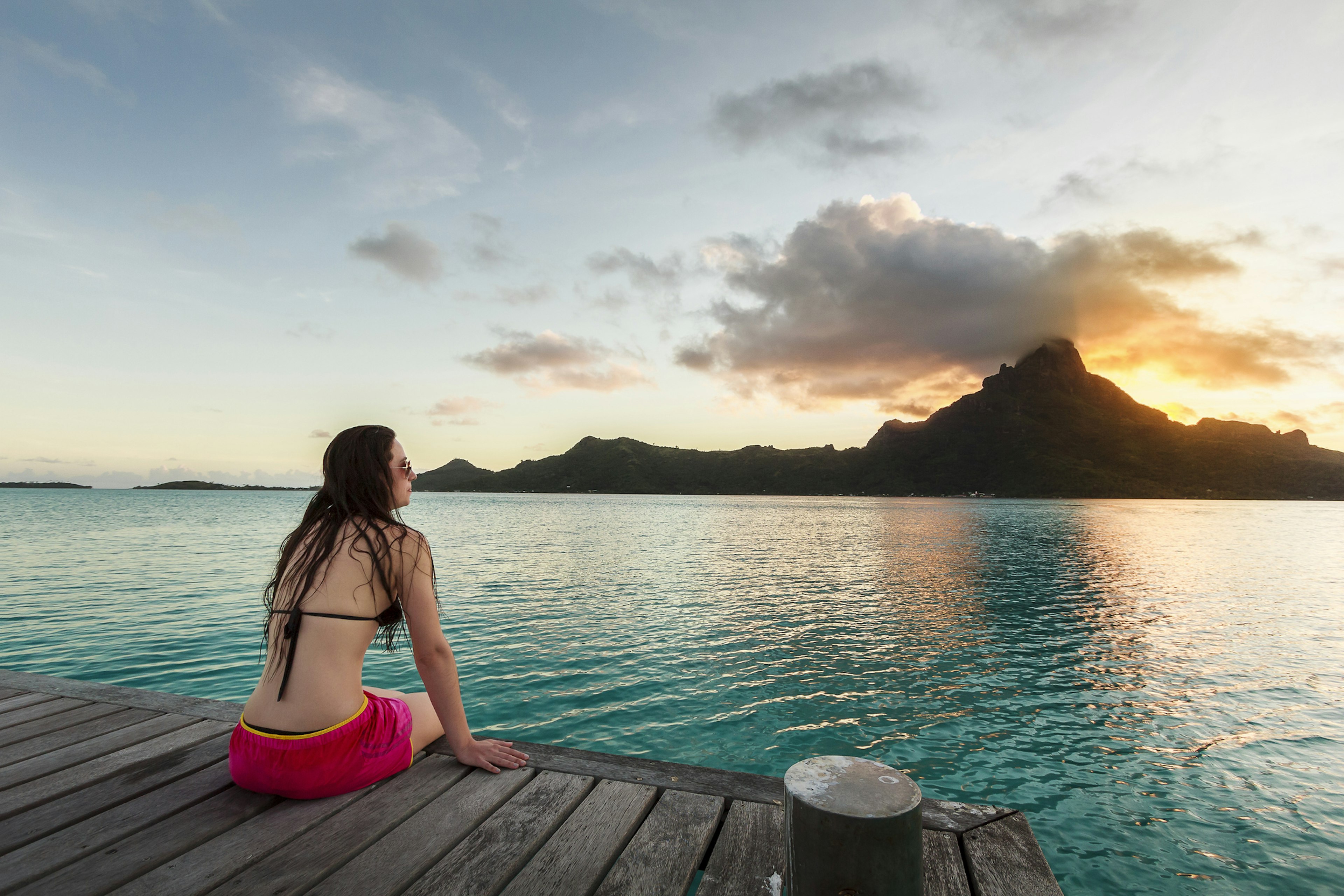 A woman is sitting on a jetty with her back to the camera watching the sunset behind Mt Otemanu in Bora Bora