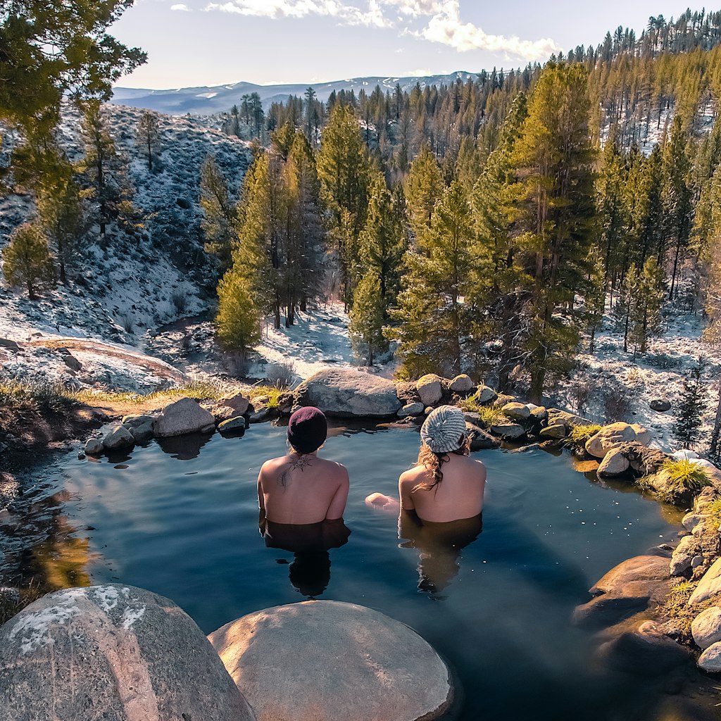 two people with winter hats sitting in a rocky pool in the mountains surrounded by snowdusted trees