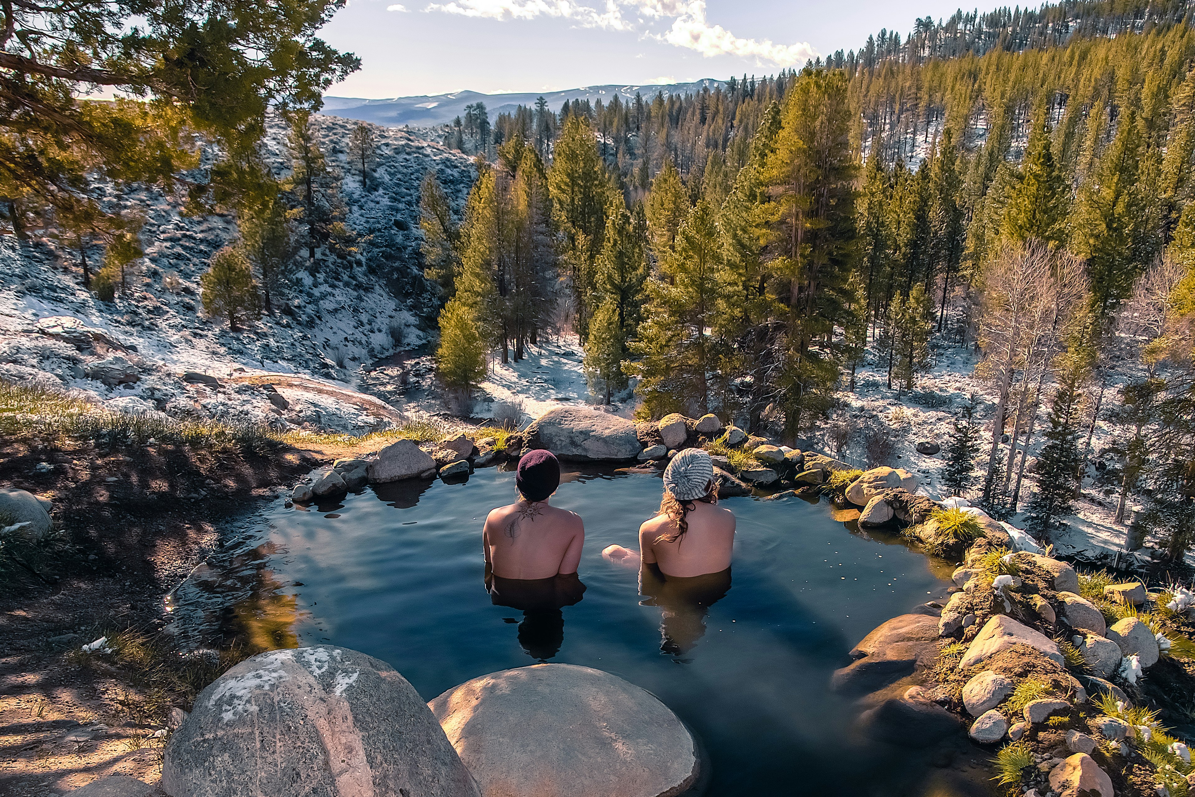 two people with winter hats sitting in a rocky pool in the mountains surrounded by snowdusted trees