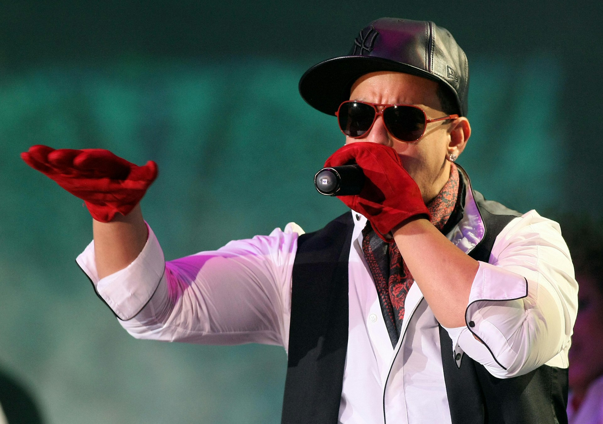 Reggaeton musician Daddy Yankee performs on stage. He's wearing red gloves, black leather baseball cap, a white button-up and black vest.  