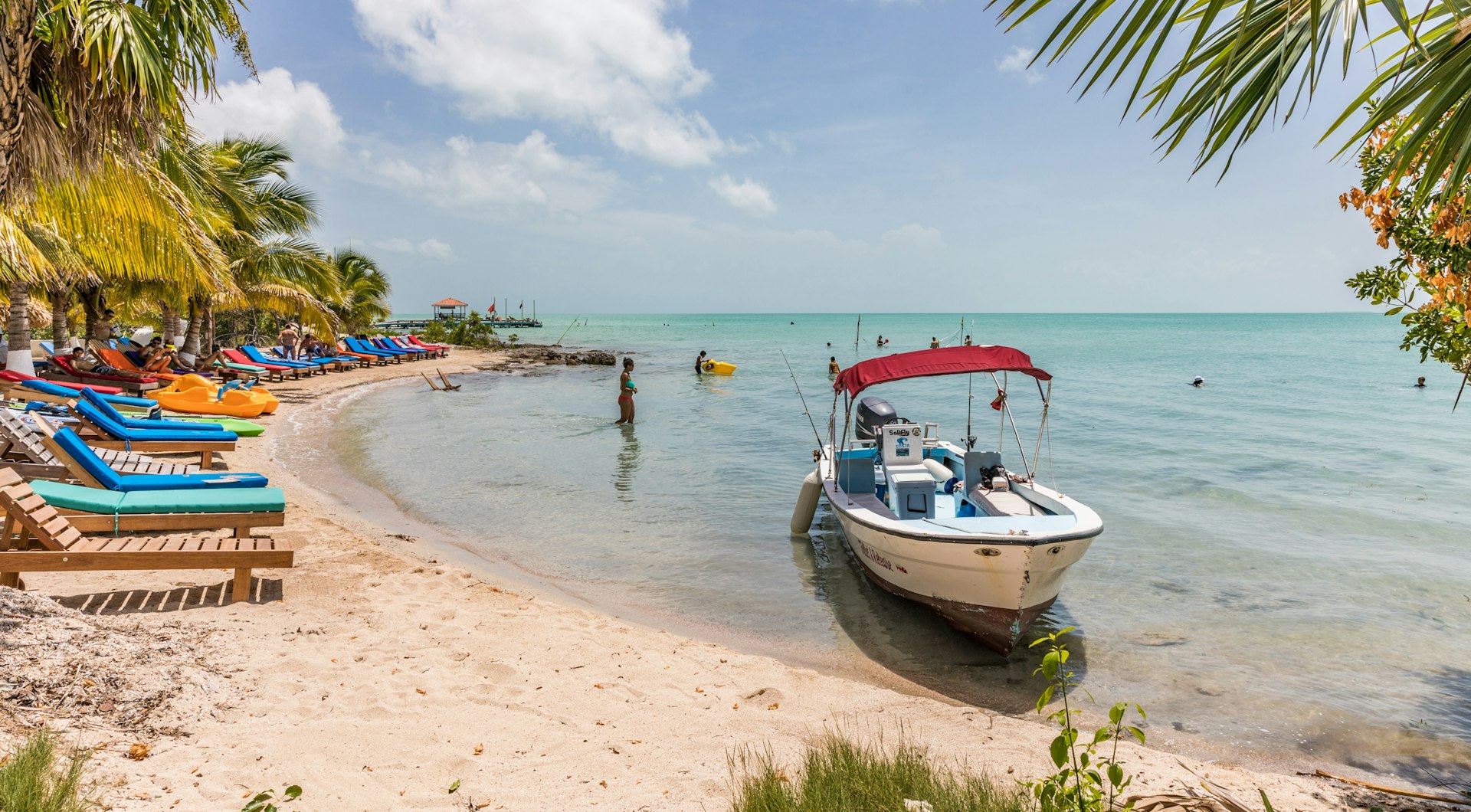 Lounge chairs, a woman in a bikini wading in the water, and a boat anchored just off the beach
