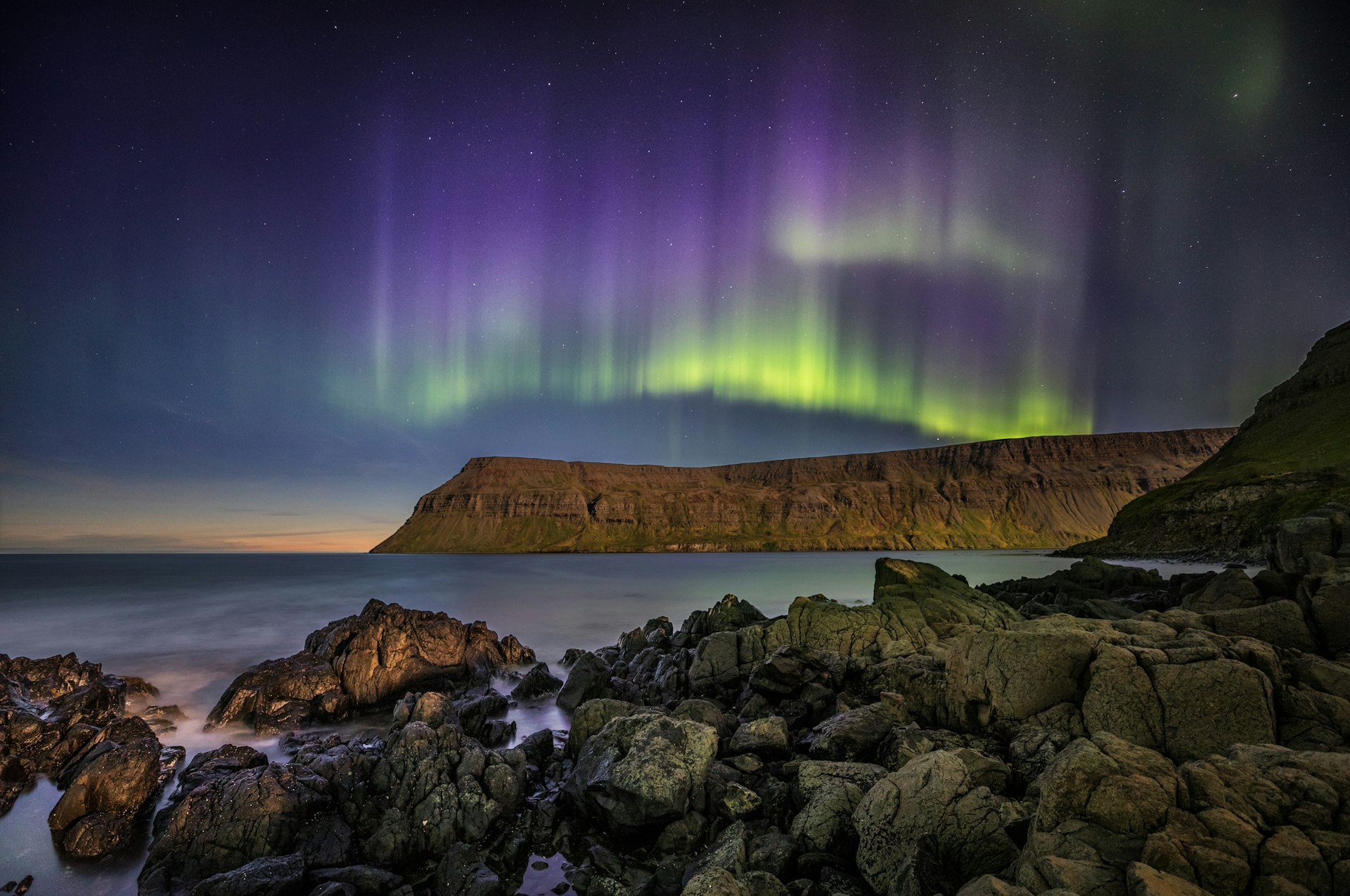 The night sky streaked with purple, green and yellow light as the Northern Lights sweep above a cliff top 