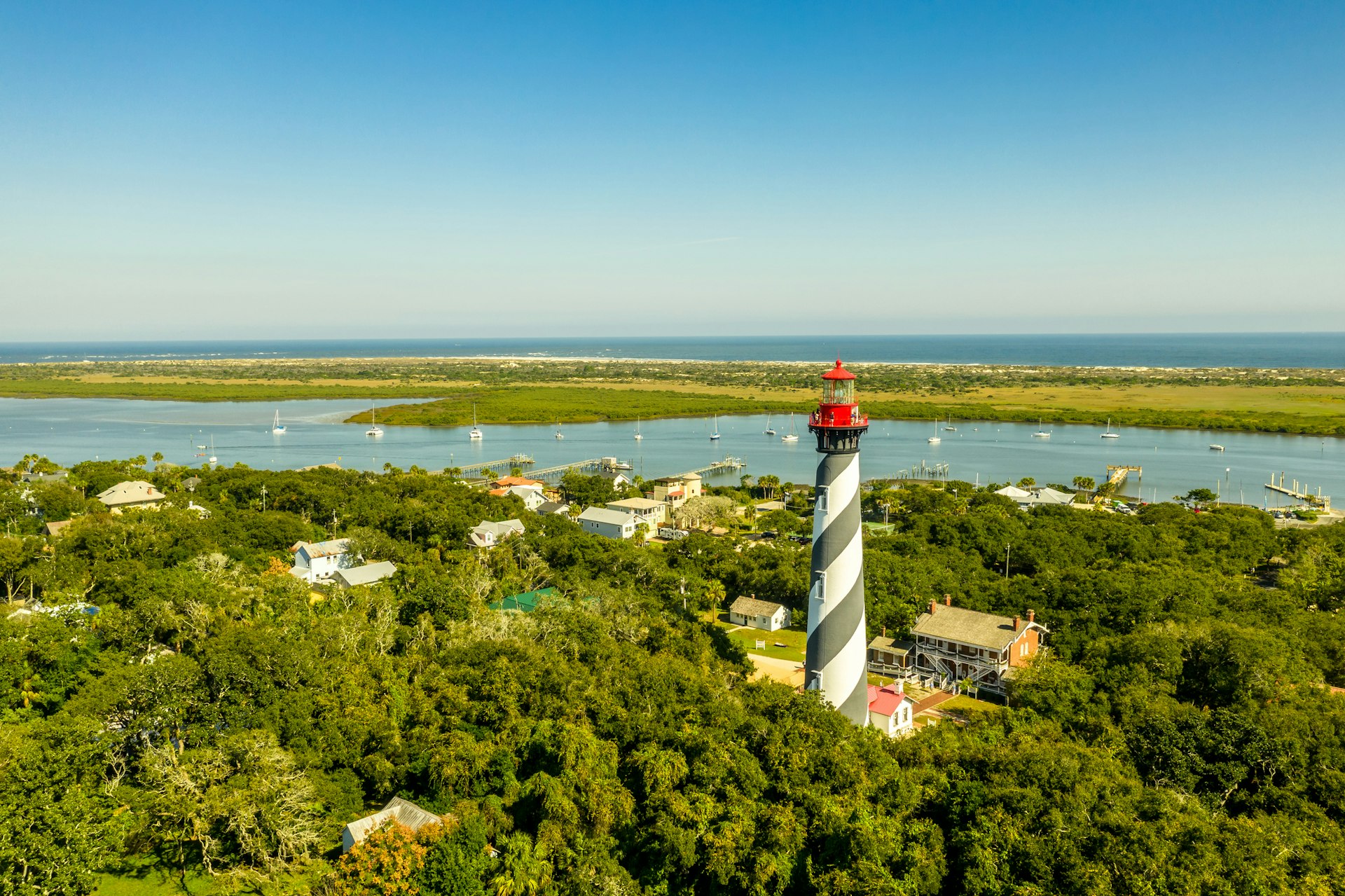 Aerial view of a black-and-white striped lighthouse. A peninsula in the distance covered in greenery gives way to a long stretch of golden sand