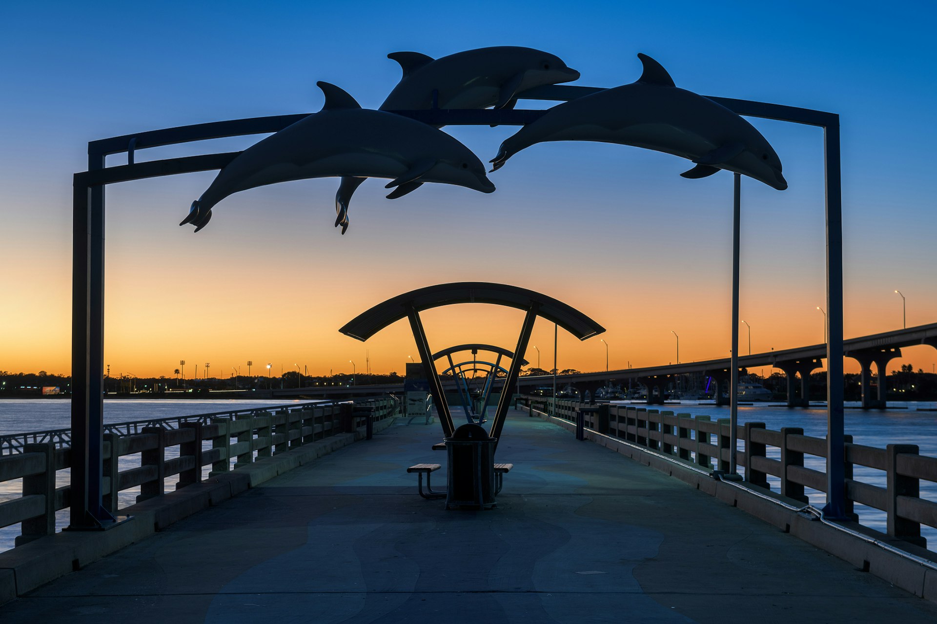 A retro pier with dolphin-shaped decorations in silhouette as the sun sets casting orange and blue light in the sky
