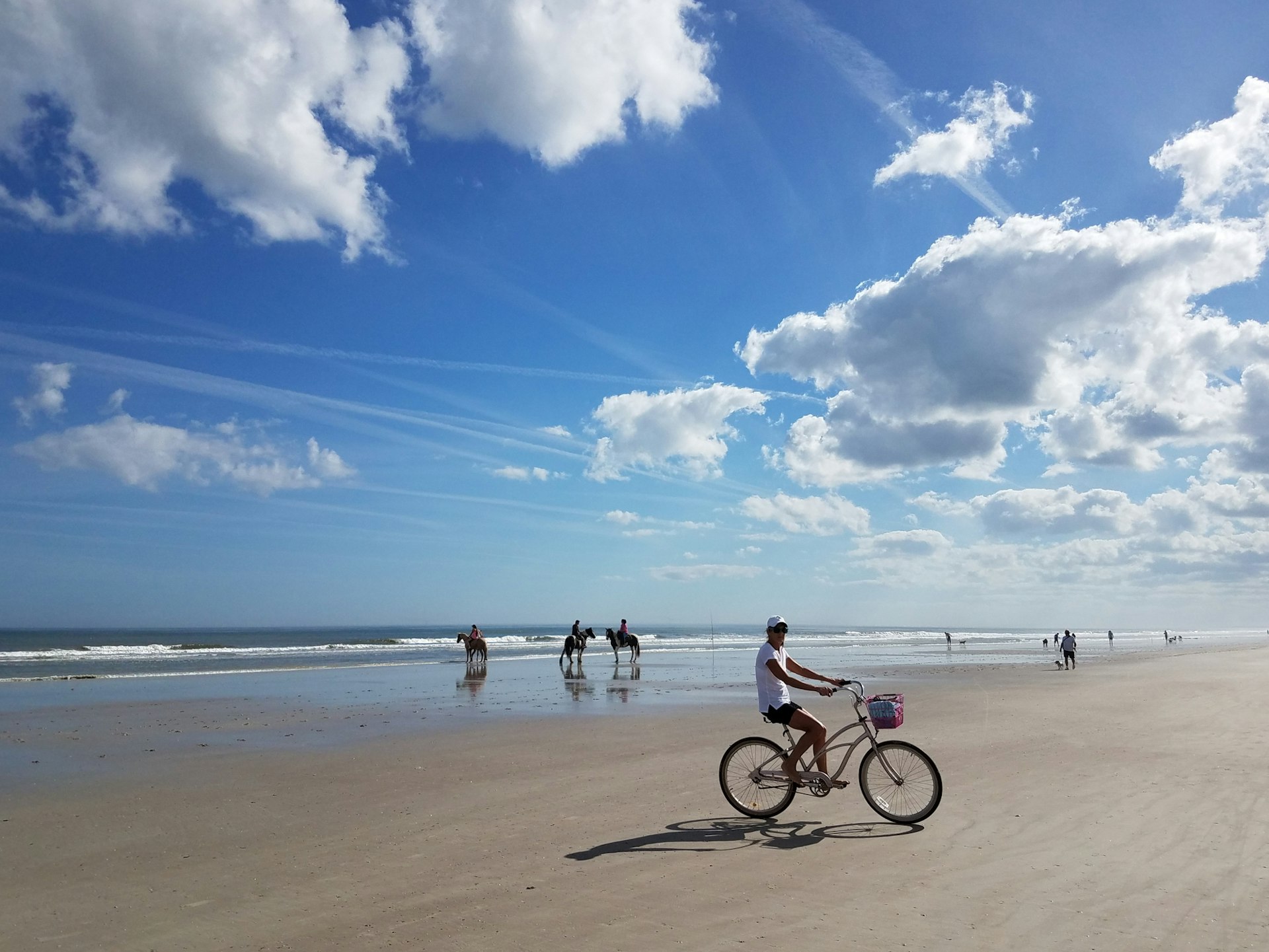 A woman rides a bicycle on a firm-sand beach. Behind her are three people on horseback