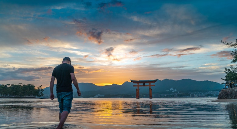 A tourist walks on the sea with the Itsukushima Shrine Torii gate in the back during sunset