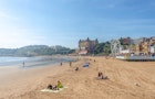 People enjoy the sunshine on the beach at Scarborough in late summer. A hotel and the the promenade line the beach. A blue sky is overhead.