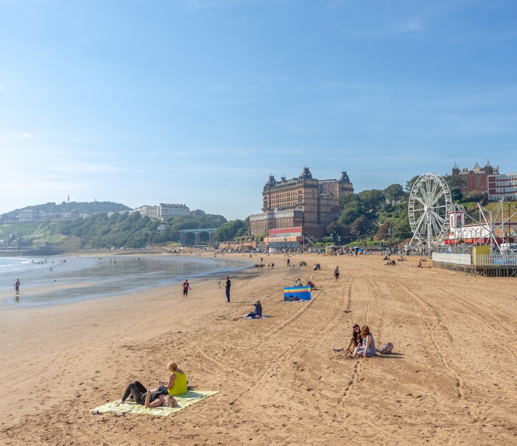 People enjoy the sunshine on the beach at Scarborough in late summer. A hotel and the the promenade line the beach. A blue sky is overhead.