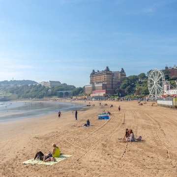 People enjoy the sunshine on the beach at Scarborough in late summer. A hotel and the the promenade line the beach. A blue sky is overhead.