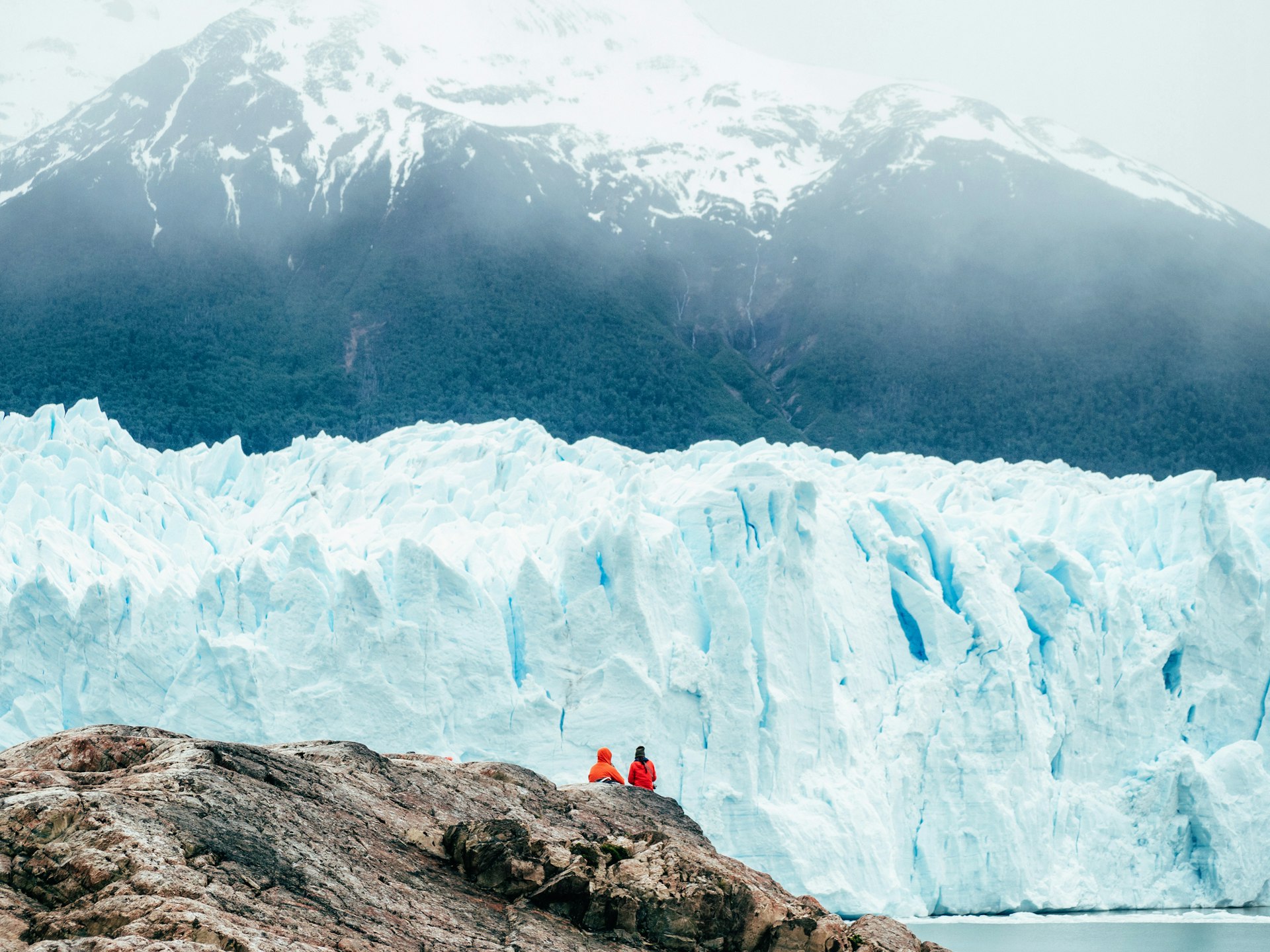 Two people sit at a viewpoint admiring a majestic blue-white wall of ice rising in front of them