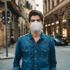 Portrait of a young adult man wearing a protective face mask and looking at camera. He's outdoor in the city. He's smiling behind the face mask. He's wearing the face mask due to the Covid-19 coronavirus pandemic.