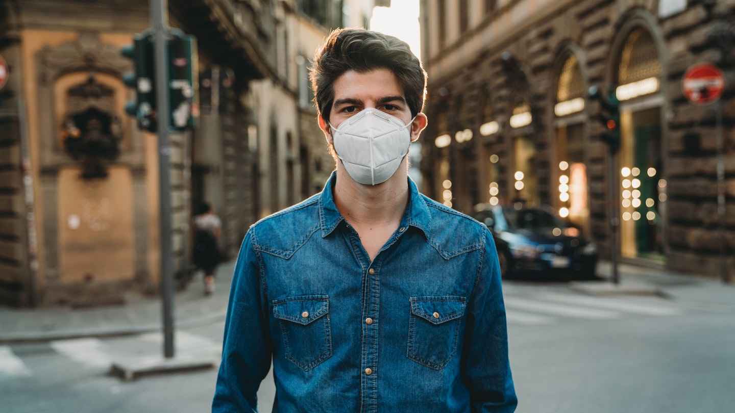 Portrait of a young adult man wearing a protective face mask and looking at camera. He's outdoor in the city. He's smiling behind the face mask. He's wearing the face mask due to the Covid-19 coronavirus pandemic.