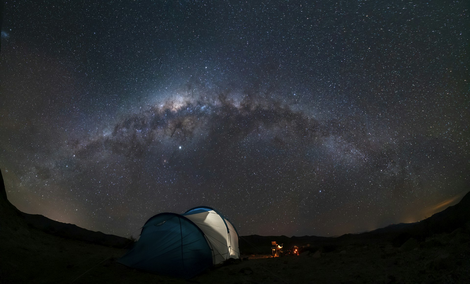 A tent, a campfire and a dark sky lit up with stars with the sweep of the Milky Way