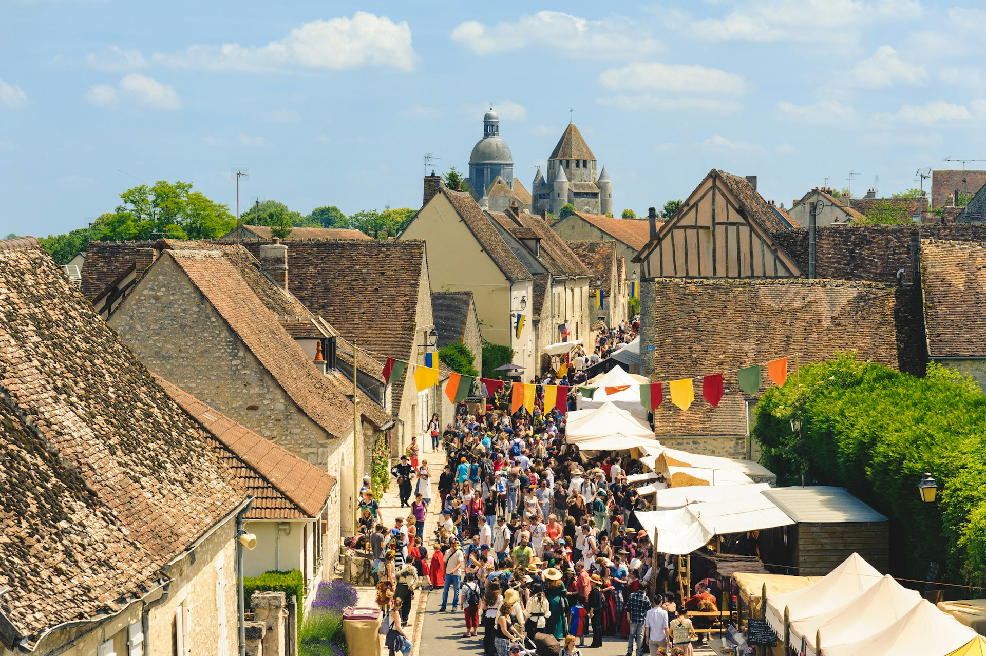 An aerial shot of a medieval town. The narrow street is decorated with colorful bunting as people attend a festival there
