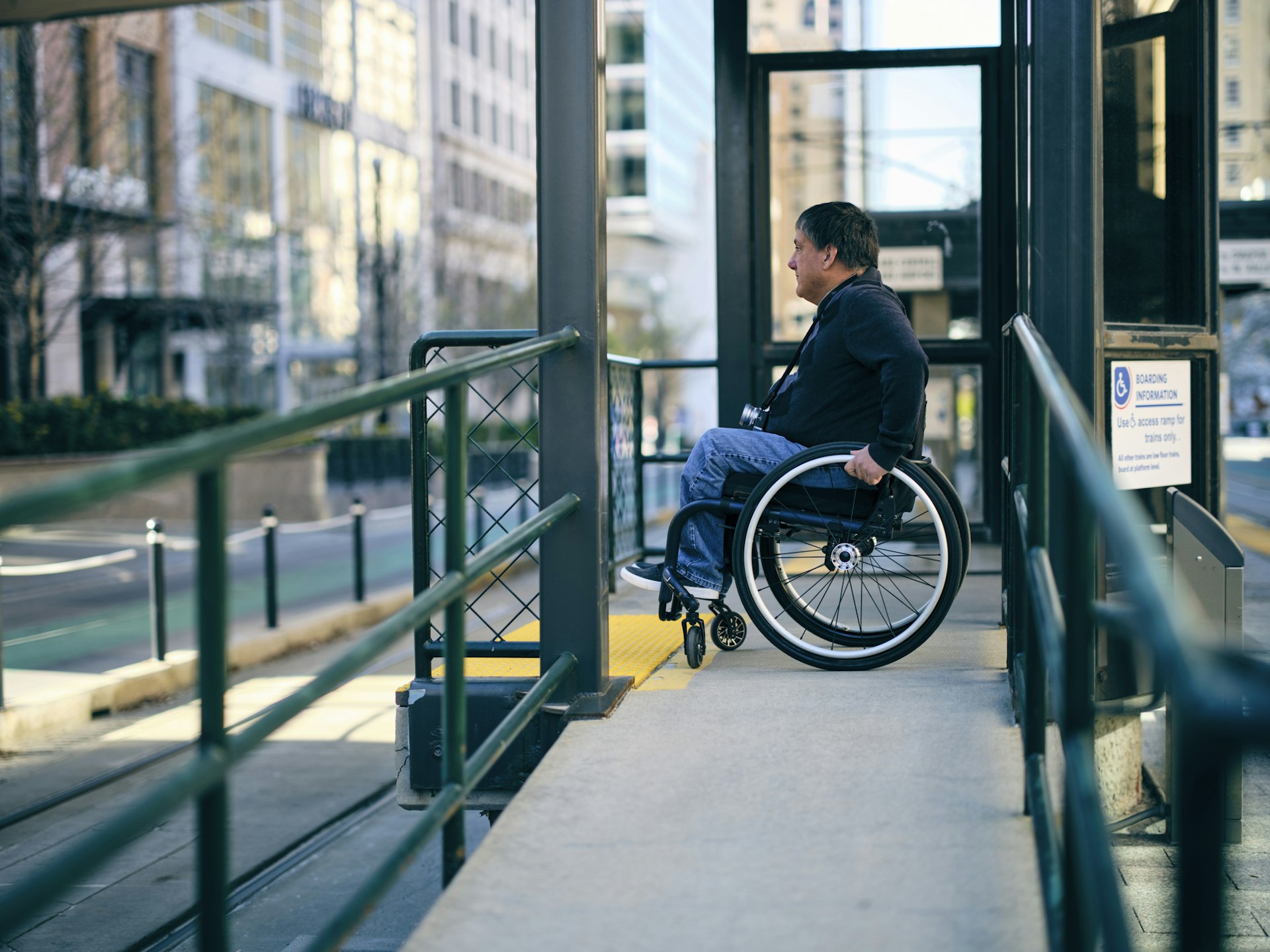 A man in a wheelchair waits on a ramp to board a light rail service in a city