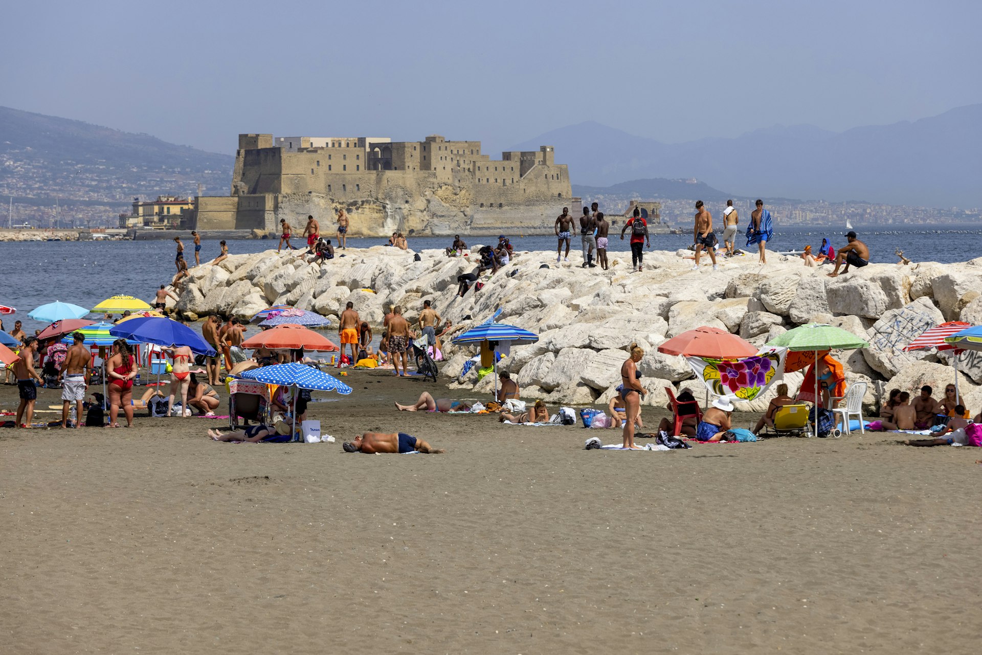 People relaxing on a beach, with colorful umbrellas and a large fortress-like building in the background