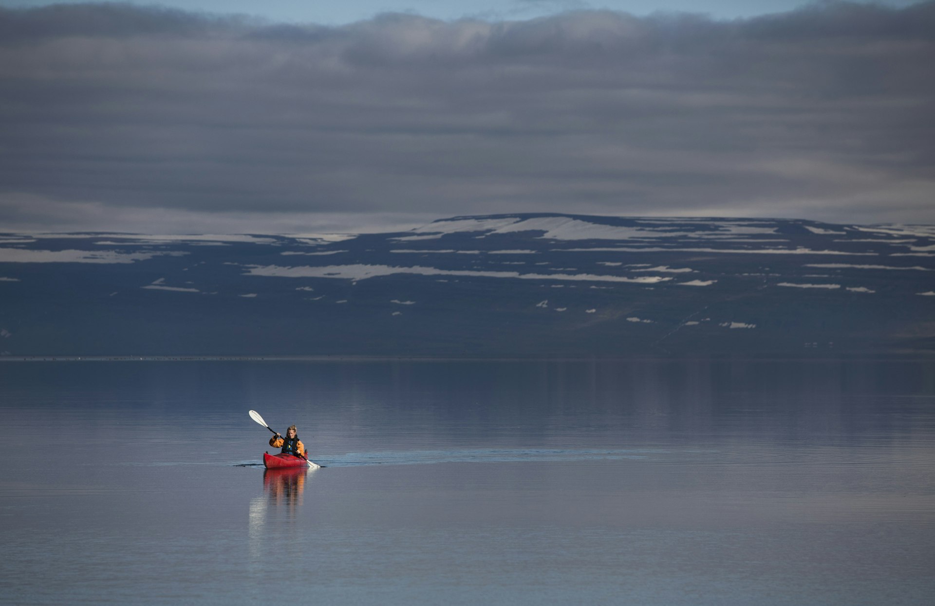 A woman in a red kayak paddles across a very still body of water with snow-capped hills in the distance