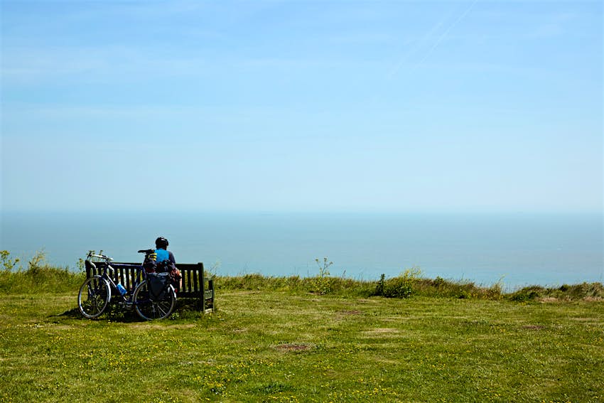 A lone cyclist sits on a bench facing the sea with a bicycle leaning against the back of the bench