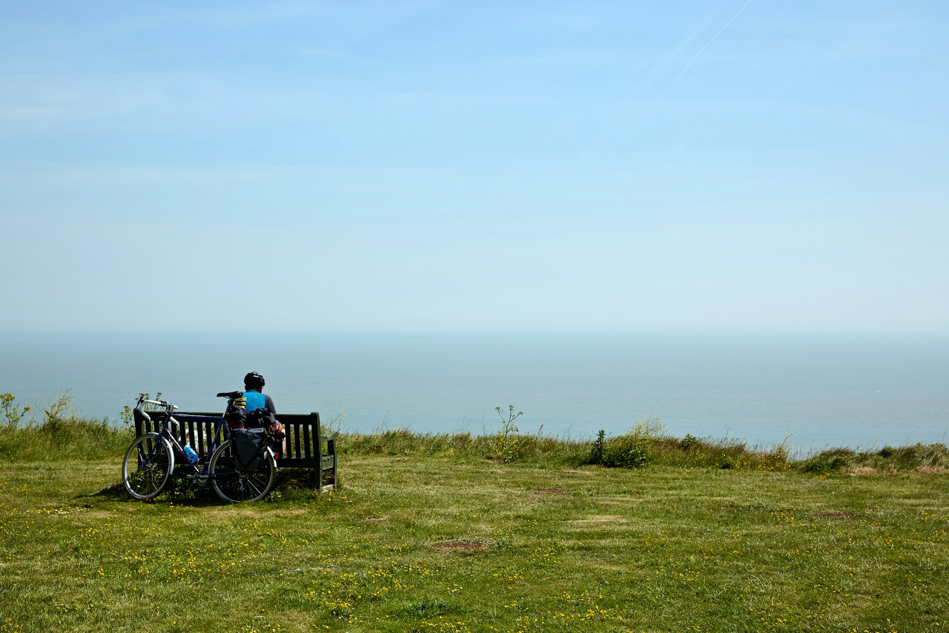 A solo cyclist sits on a bench looking out to sea with a bike leaning against the back of the bench