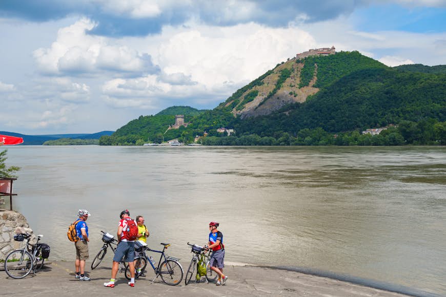 A group of cyclists pause with their bikes on a stretch of sand at the edge of a river, overlooked by a castle on a cliff