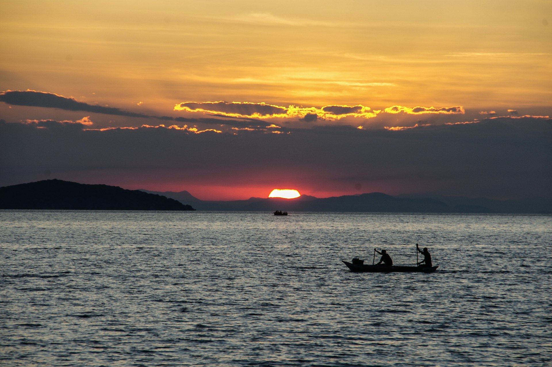 Two fishers in a dugout canoe are paddling on a lake as the sun sets, covering the sky in orange and purple