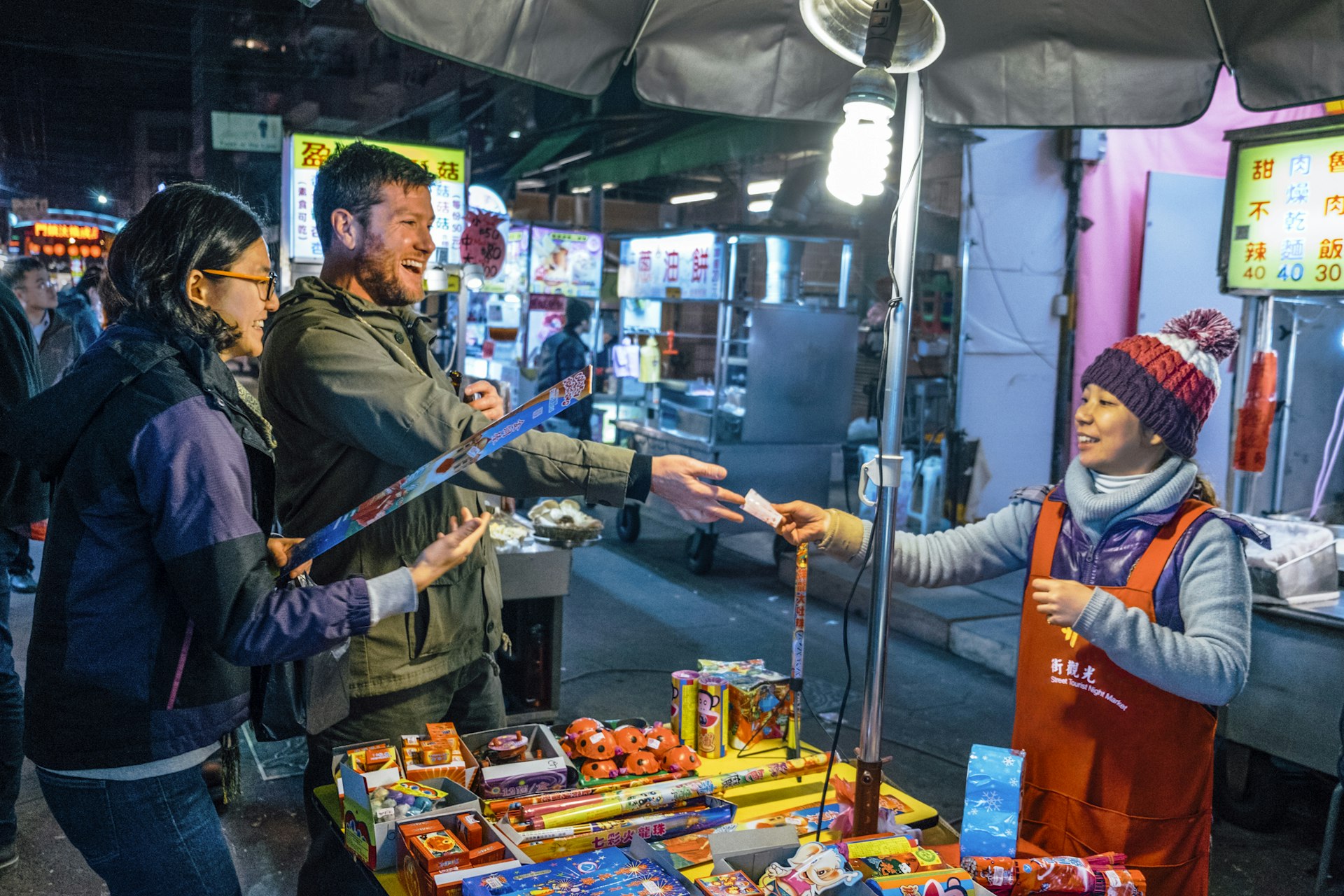 Two people smile as they hand over money to a vendor at a night market stall 