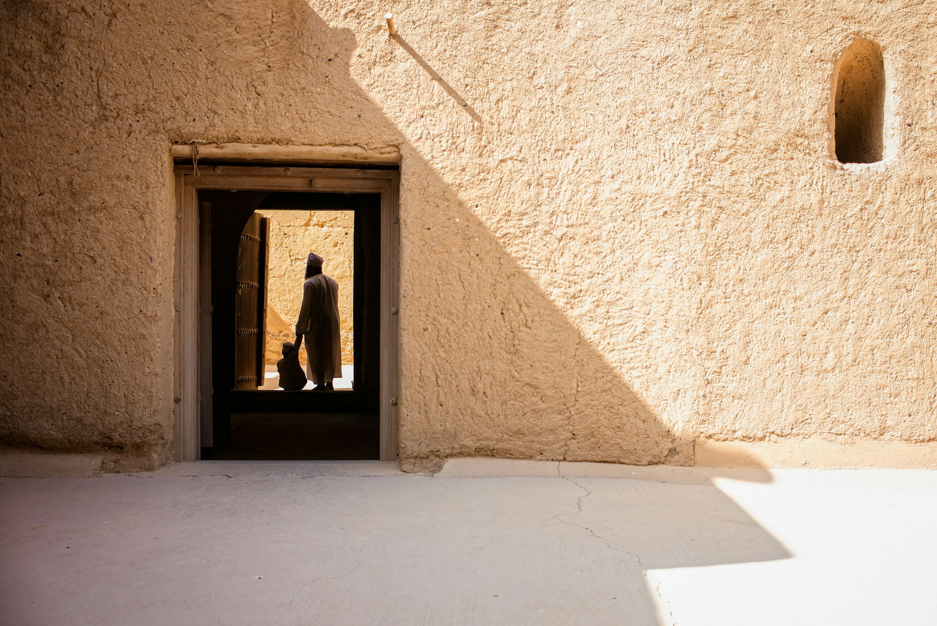 A man and a child holding hands in silhouette are framed by a doorway in a large sand-colored fortress building