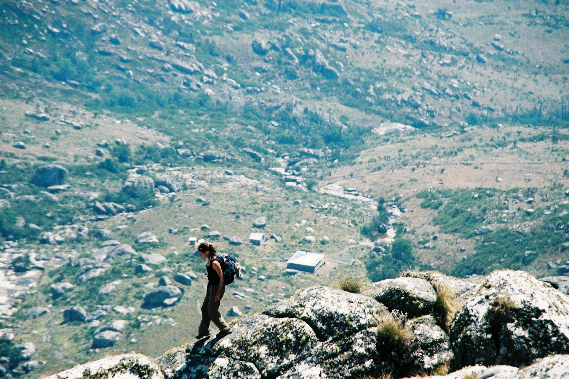 A solo woman carrying a backpack hikes down a rocky mountain ridge. A hut is in the valley below.