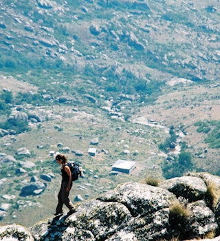 Side view of a Female hiker walking down on a mountain ridge with a day pack and the Chisepo Hut in the background below Mount Mulanje Mountain Range Mulanje Zoma Plateau Malawi Africa