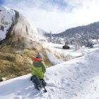 Wyoming, USA - February 21, 2018: A boy in a green jacket and snowshoes sits on a snowy back in front of Orange Spring Mound on the Upper Terraces waiting for a bison to move away from the trees at the next bend in the trail on a guided excursion around Mammoth Hot Springs at Yellowstone National Park during winter