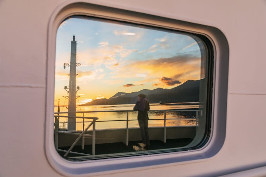 A woman is standing on the deck of a ship at sunrise looking toward the Beagle channel reflected in a window of the ship
