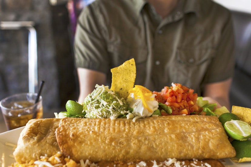 A man sitting at a Mexican food restaurant about to eat a giant chimichanga (fried burrito).