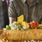 A man sitting at a Mexican food restaurant about to eat a giant chimichanga (fried burrito).