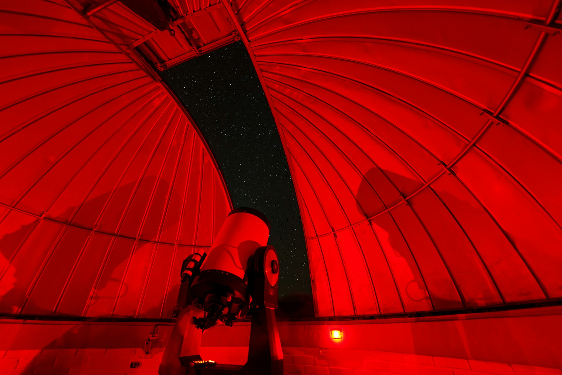 An observatory at night, lit with dim red light. A large telescope stands in the center of the observatory pointing towards the sky