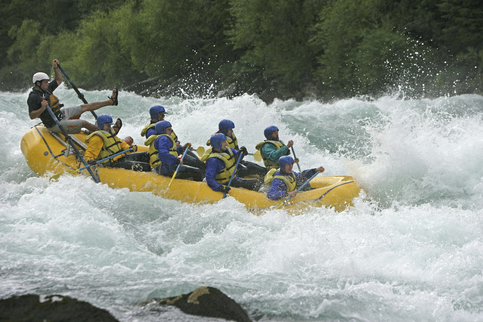 A group of eight people in blue wetsuits, helmets and yellow lifejackets white-water raft along the Futaleufu River with the leader at the back almost being chucked from his seat