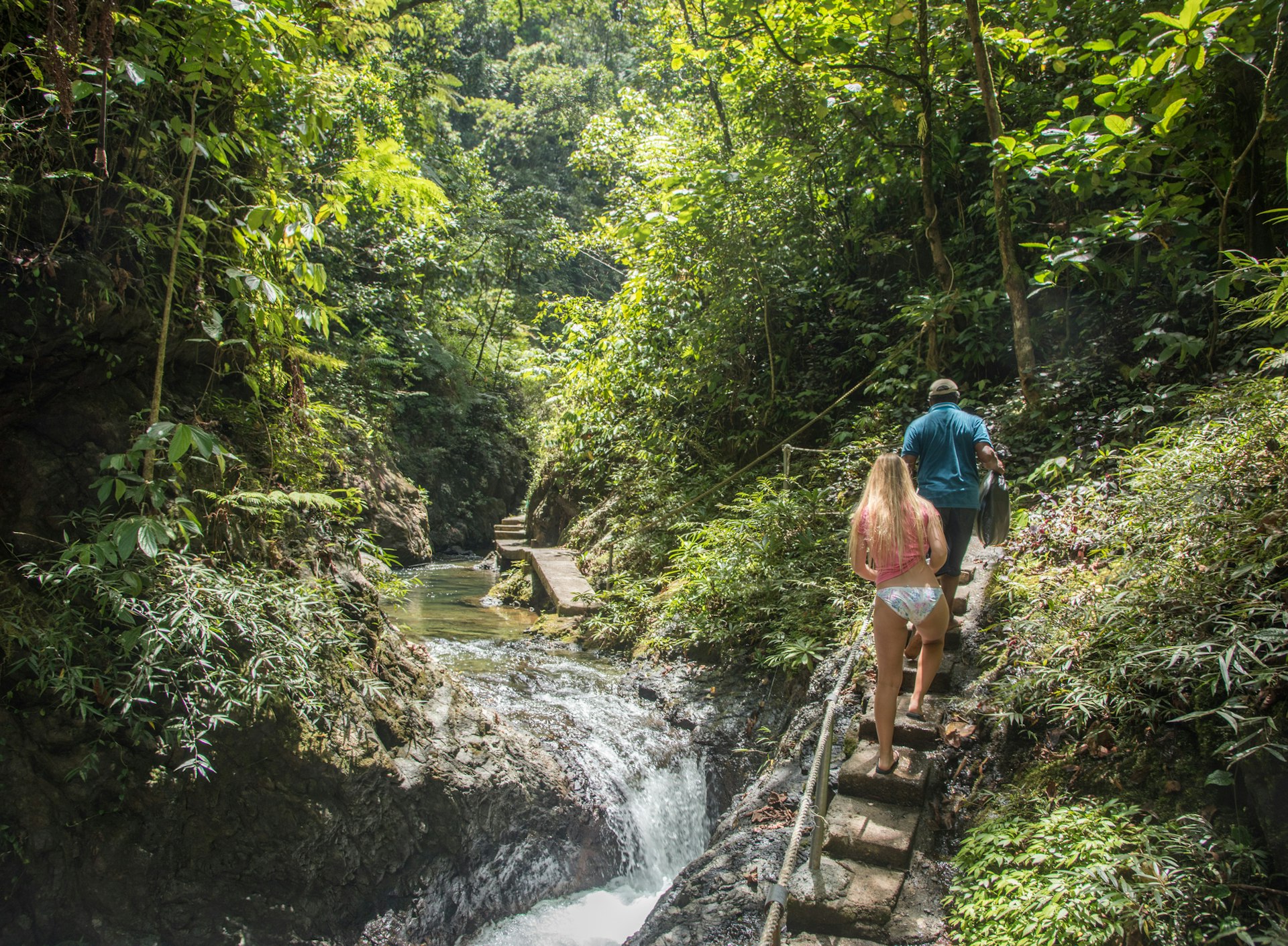 A man and woman walking along a path through the Suva rainforest in Fiji with a river flowing next to them