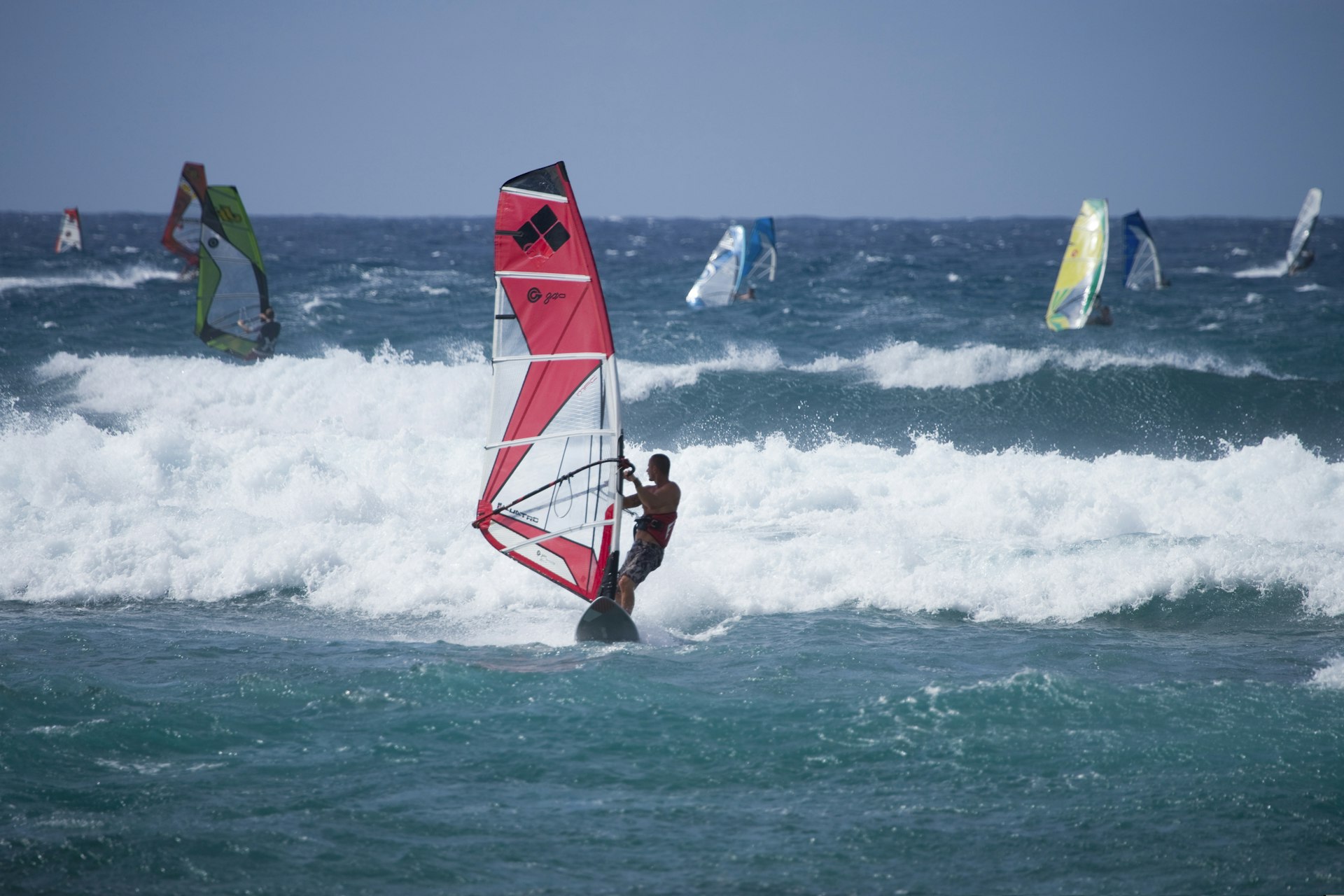 A windsurfer with a red sail attached to a board riding waves. Several other windsurfers are further out in the occean