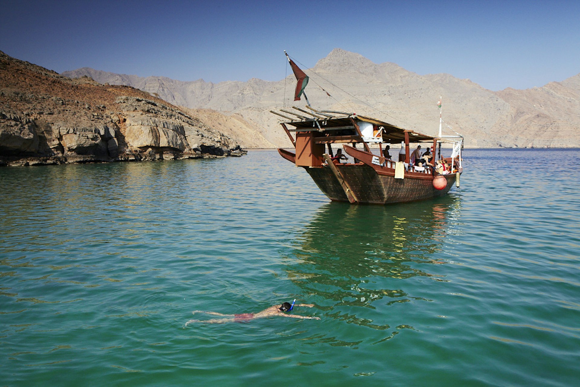 A man snorkels in front of a large wooden tourist boat in turquoise waters surrounded by sand-colored cliffs