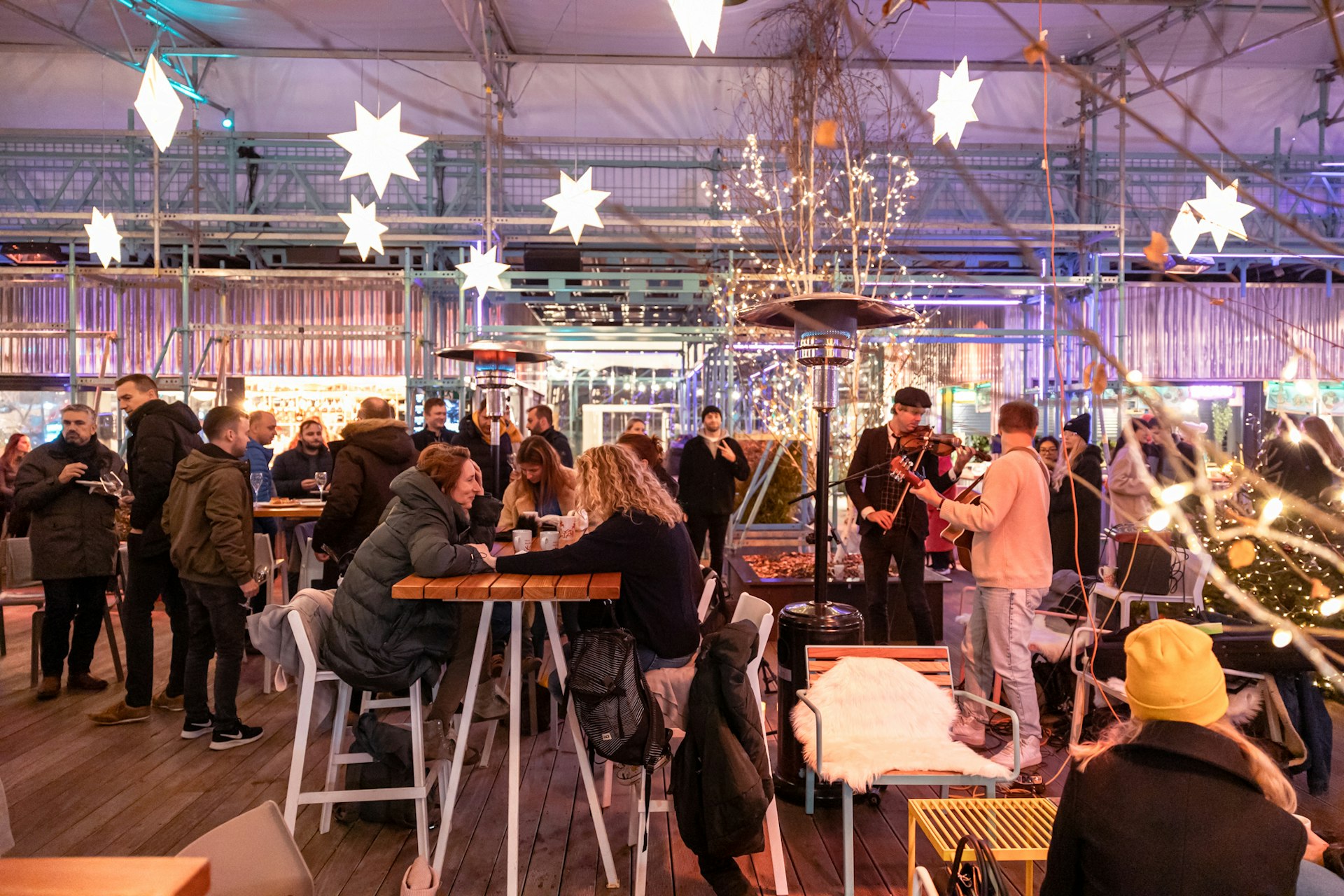 A crowd of people sit at tables in winter coats and hats at Prague's Manifesto Market Anděl with a band playing to the right hand side. The outdoor market is decorated with lights shaped as stars