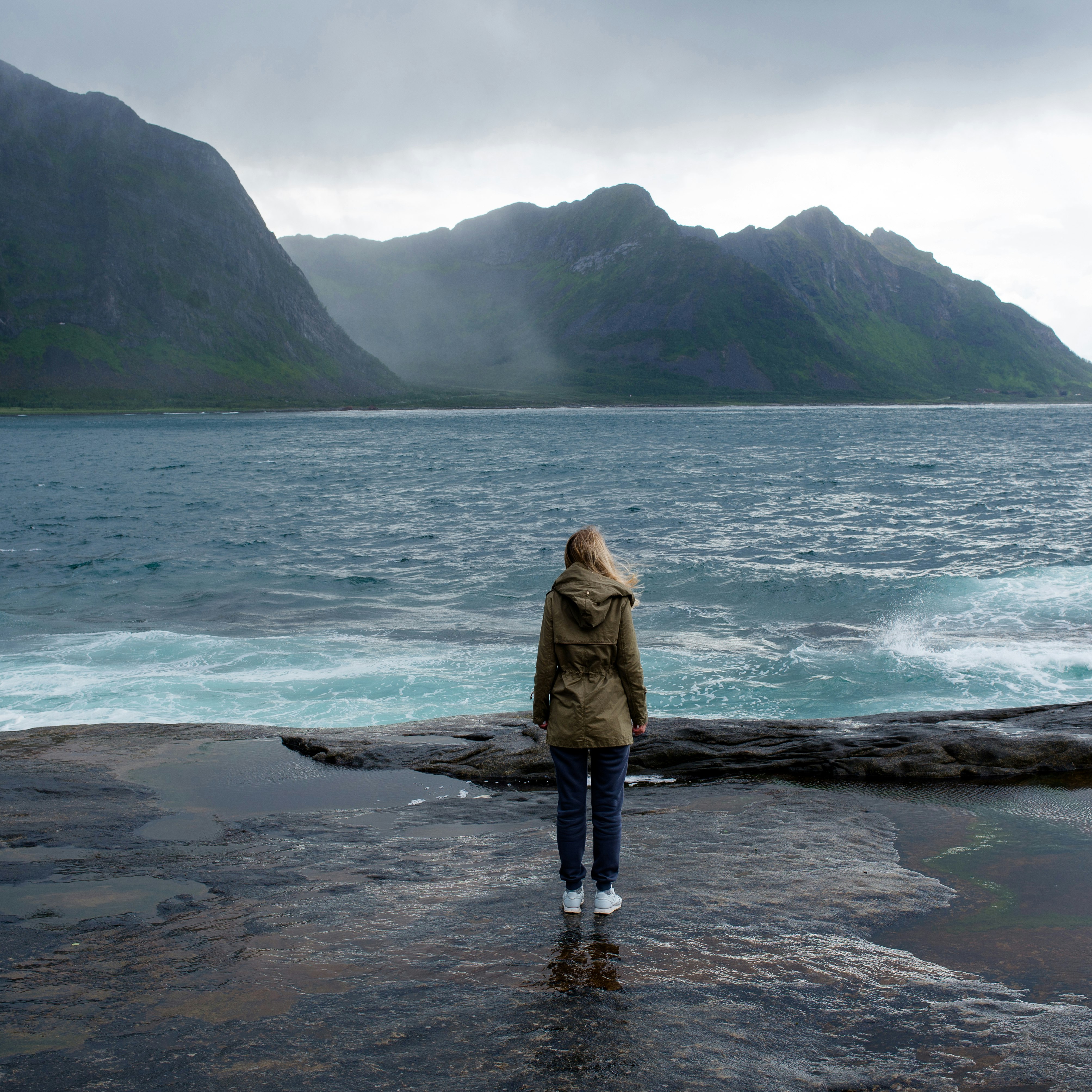 Blond hair girl with a backpack stands on big stones near the water and looks at the ocean. Waves, splashing. Enjoy the moment, relaxation. Wanderlust. Travel, adventure, lifestyle. Explore Norway