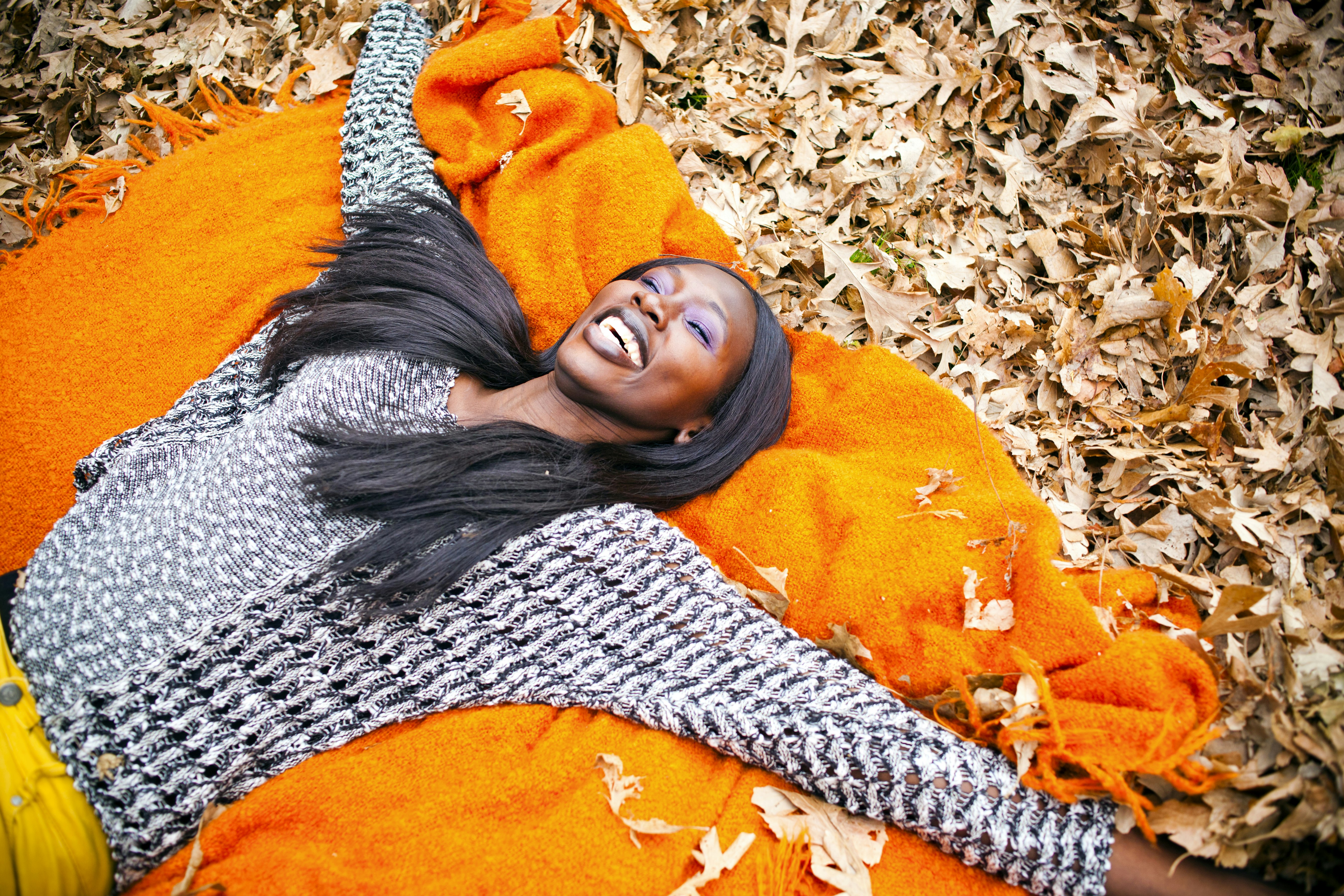 A woman in a black and white sweater and yellow pants lays on a bright orange blanket. There is a pile of leaves beneath her. 
