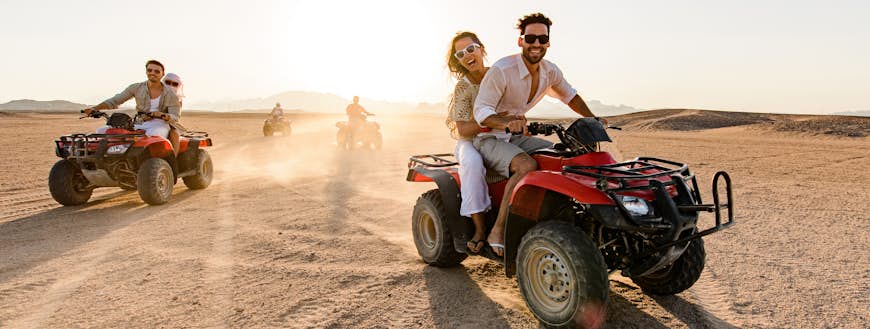 Happy friends driving quads in the desert at sunset.