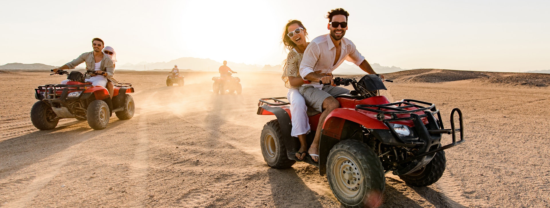 Happy friends driving quads in the desert at sunset.