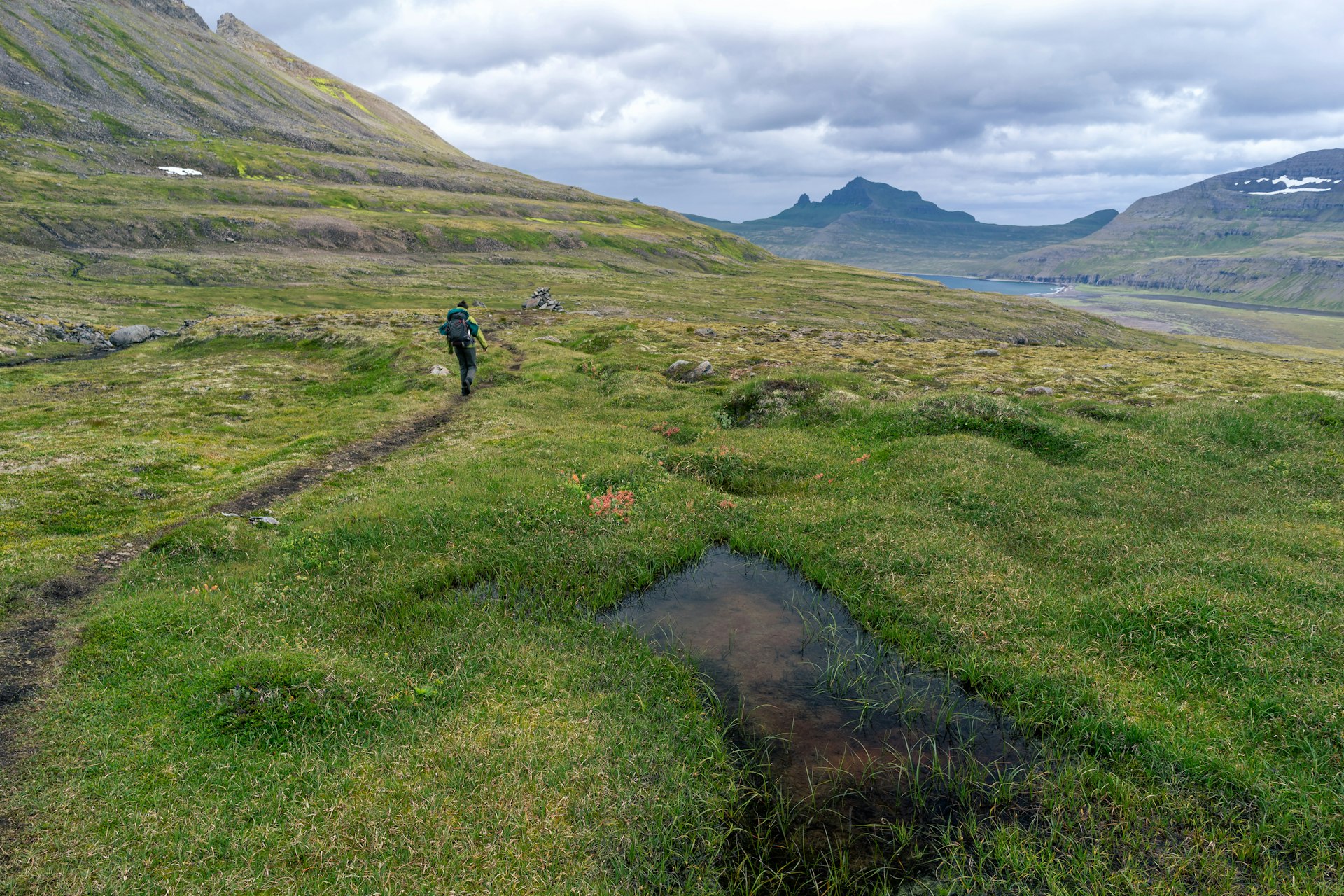 A hiker on a narrow path through a grassy valley, with clouds overhead