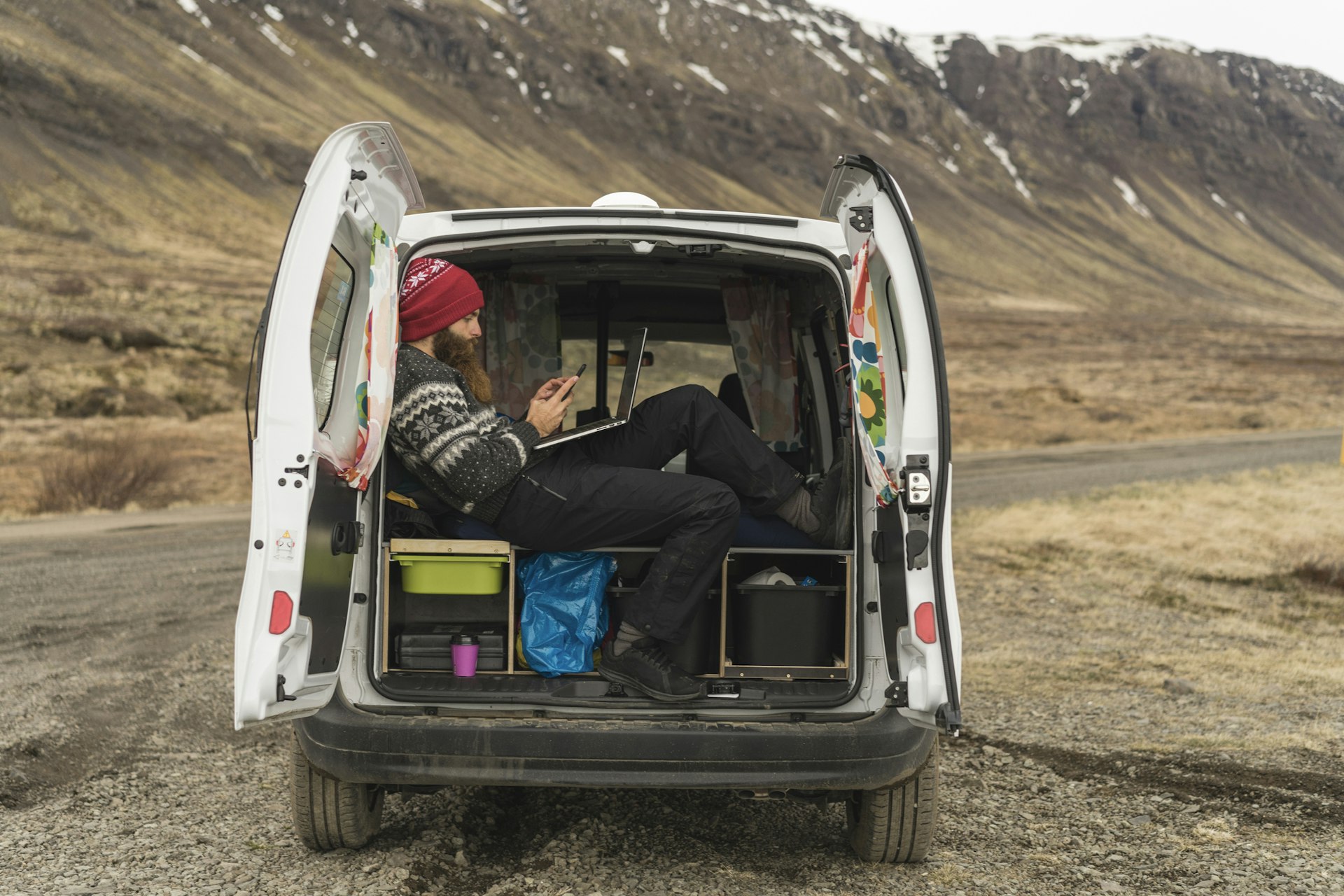 Iceland, man sitting in van using smartphone and laptop