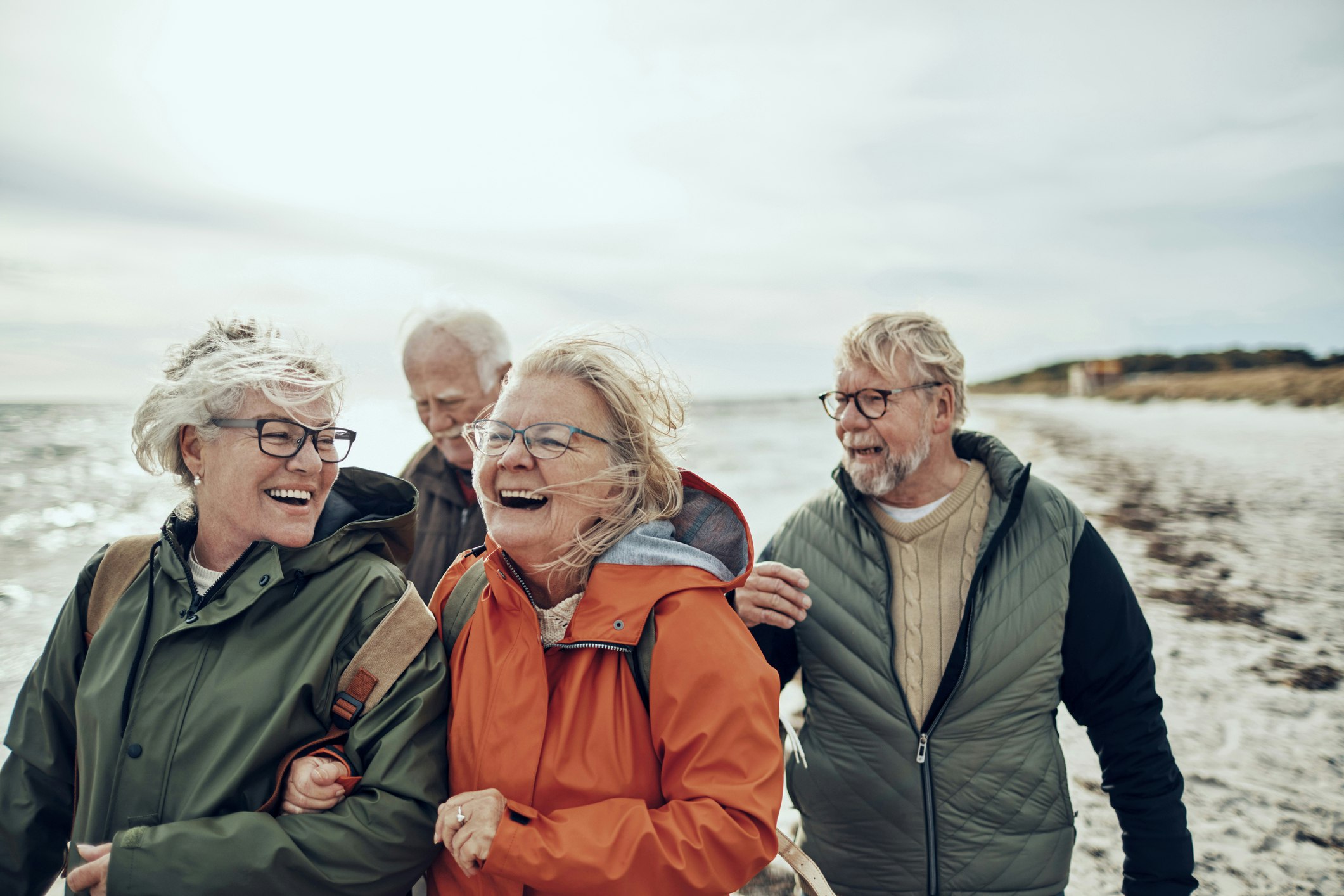 An older friend group of two men and two women walking along the beach in rain jackets while laughing together