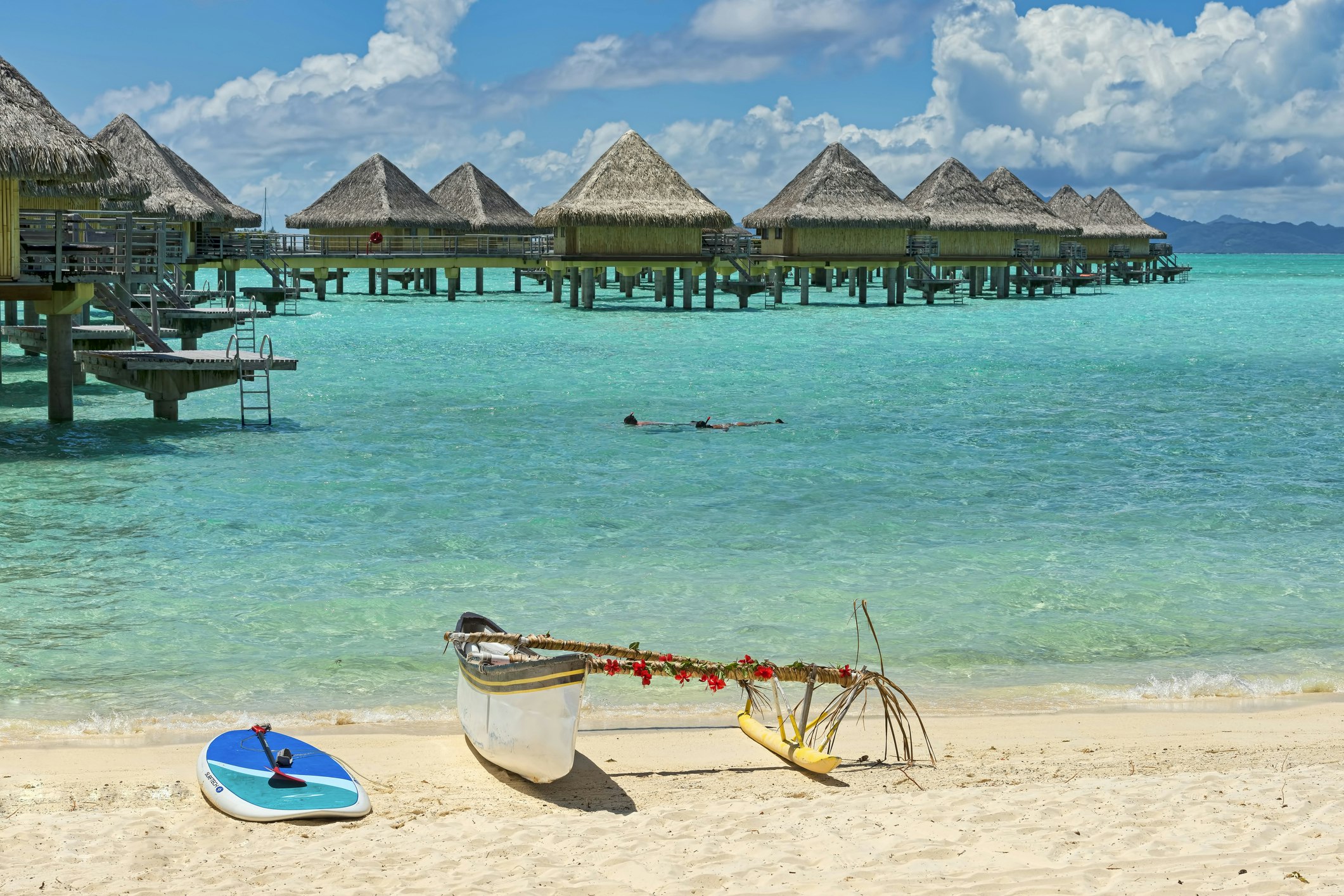 A decorated outrigger boat rests on the beach in front of overwater bungalows in Bora Bora