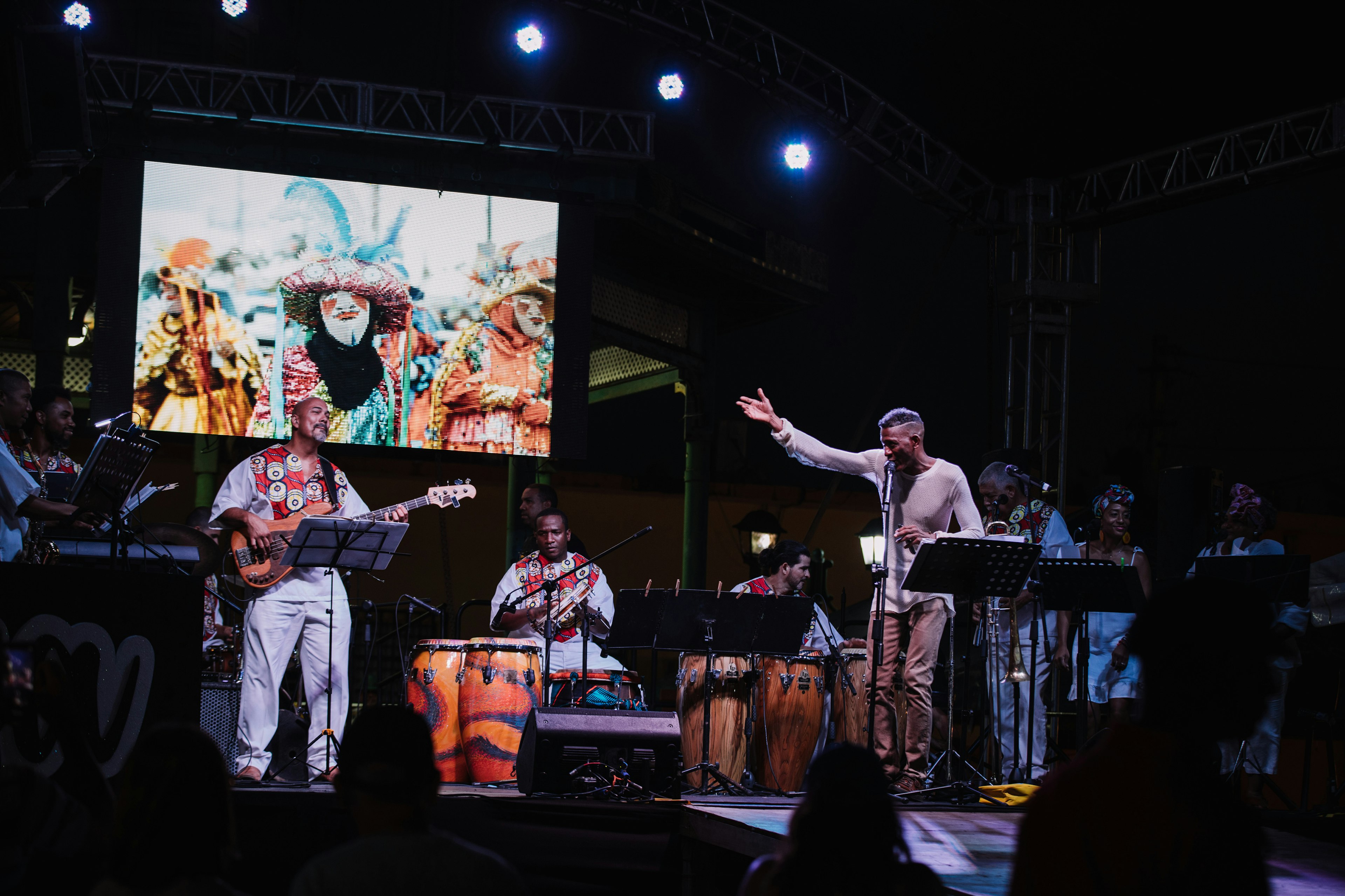 A plena band performs on stage; to the right is a man standing in front of a microphone gesturing to a video screen behind the band.